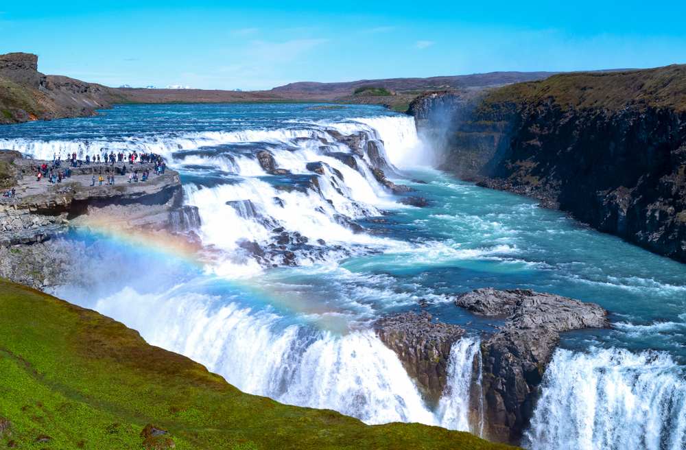 Gullfoss Waterfall in Iceland with turquoise Hvítá River, misty cascades, viewing platform tourists and rainbow over rocky canyon