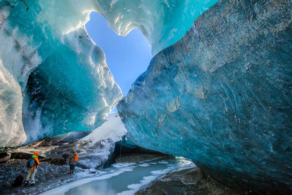 Travelers exploring a glowing blue ice cave beneath Vatnajokull Glacier, with frozen pools and sculpted ice walls in November Iceland