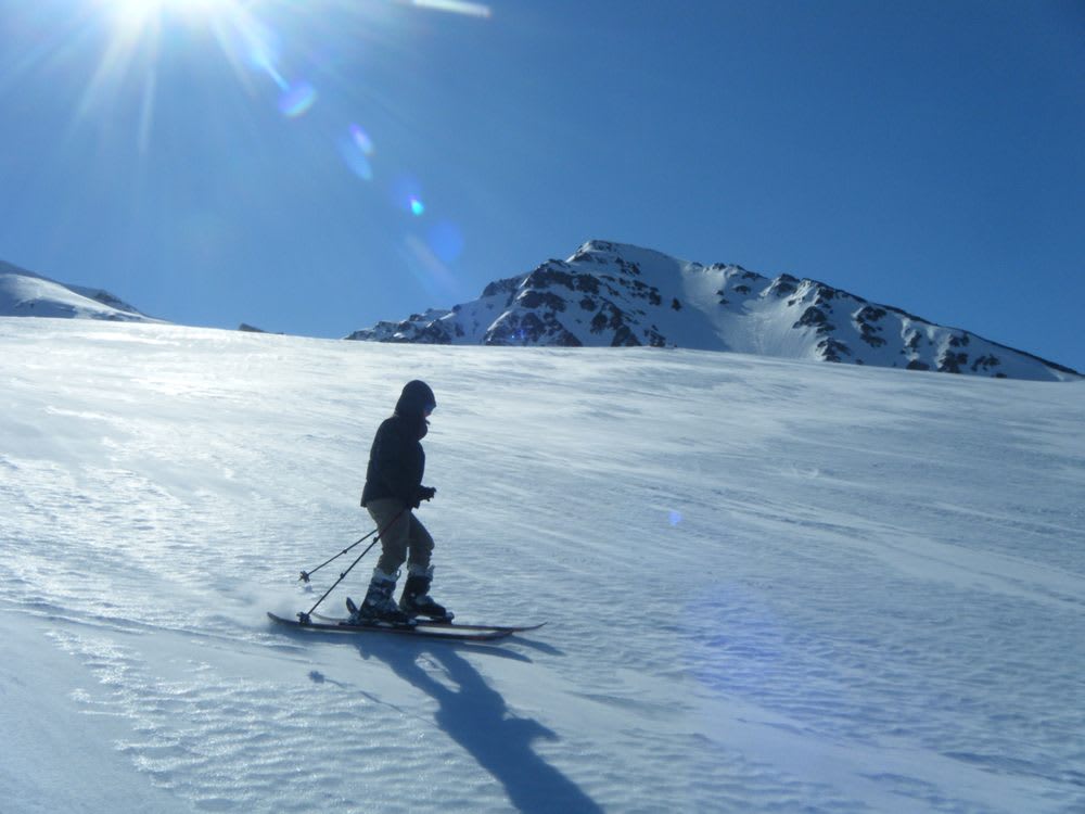 Skier gliding across a snowy Icelandic mountain slope under bright winter sun, with rugged peaks and clear blue skies in November