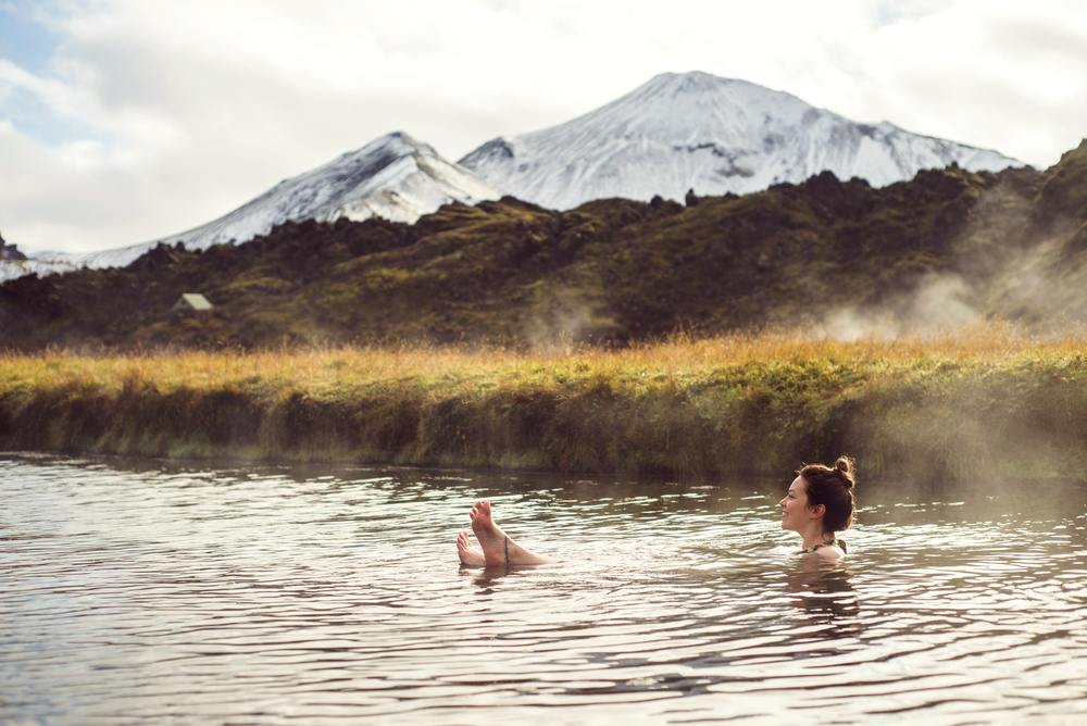 Traveler relaxing in a steaming Icelandic hot spring, with snow-dusted mountains and volcanic fields behind her on a November wellness trip