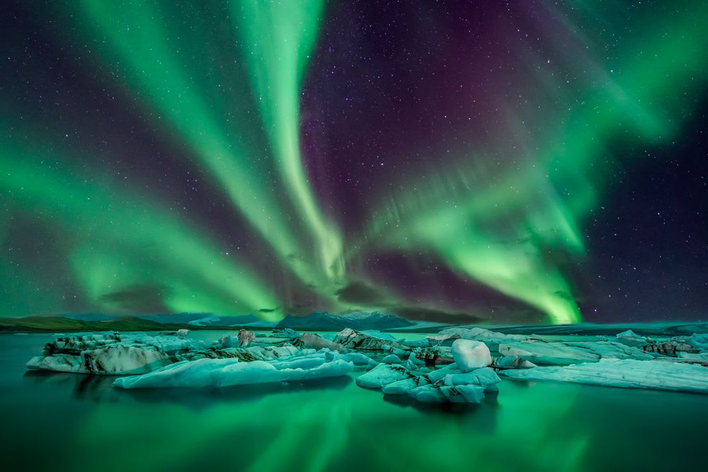Vivid green northern lights over Iceland’s glacier lagoon and floating icebergs, highlighting winter as a magical time to visit Iceland