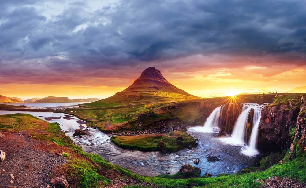 Kirkjufell mountain and waterfalls at fiery fall sunset in Iceland, with dramatic clouds and green hills for September to November travel