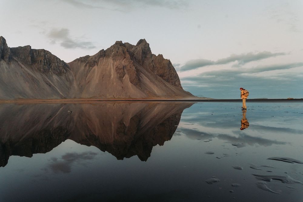 Hiker reflected on wet Stokksnes black sand beach with Vestrahorn mountains under moody October skies in southeast Iceland travel guide