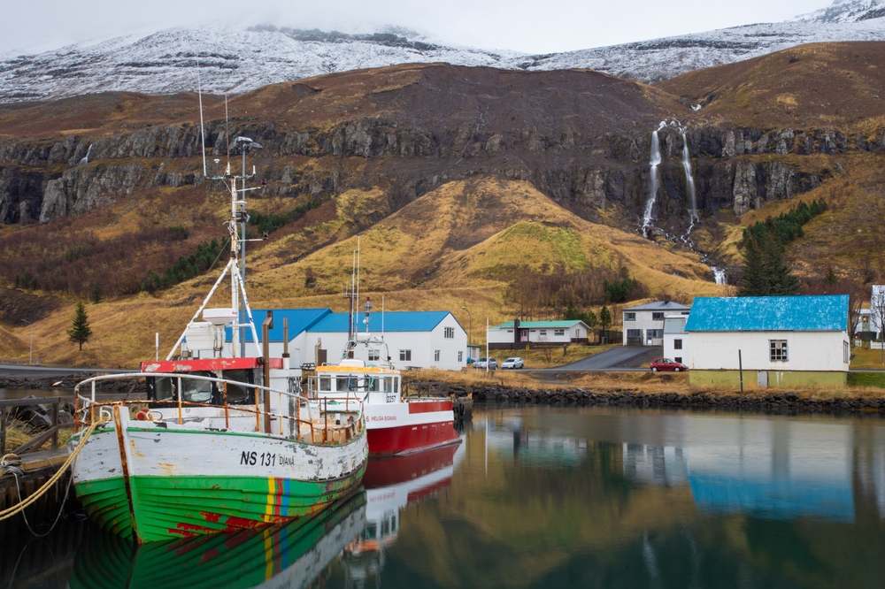Fishing boats reflected in a calm East Iceland fjord harbor below blue-roofed homes, waterfalls, and snow-dusted October mountains