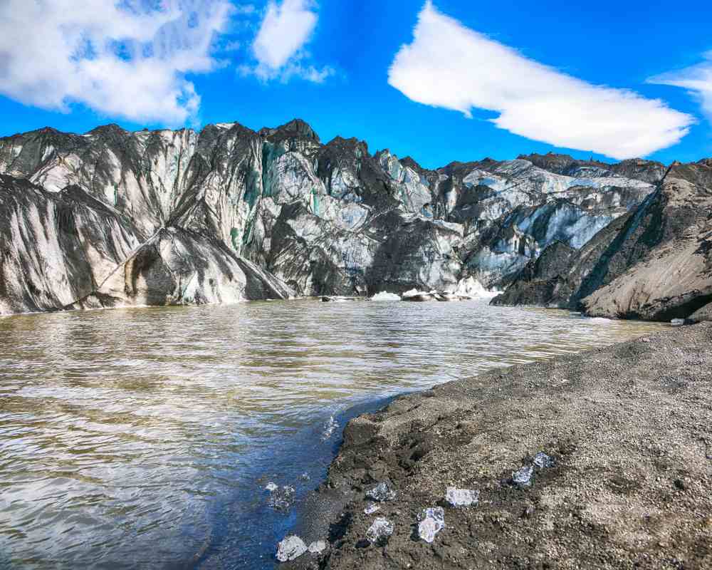 Glacier lagoon with black volcanic shore, scattered ice and rugged glacier mountains under blue September skies in Iceland’s highlands.