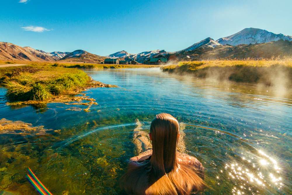 Traveler soaking in a steaming natural geothermal pool, with clear blue water, golden grass and mountain views in Iceland’s September sun.