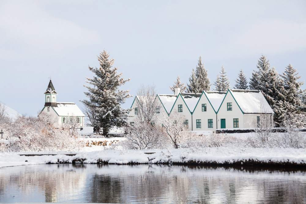 Snow-covered Þingvellir Church and historic farmhouses reflected in a calm river, surrounded by frosty trees in Iceland in winter