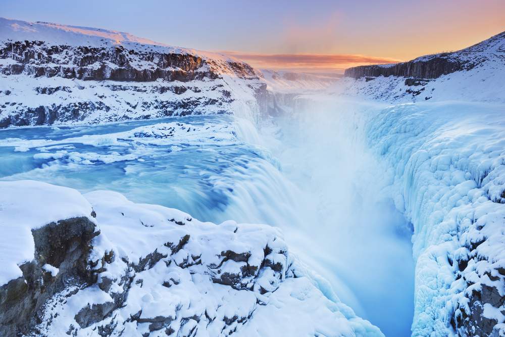 Frozen Gullfoss waterfall plunges through a snowy canyon at sunset, with icy cliffs and misty blue water on Iceland’s Golden Circle