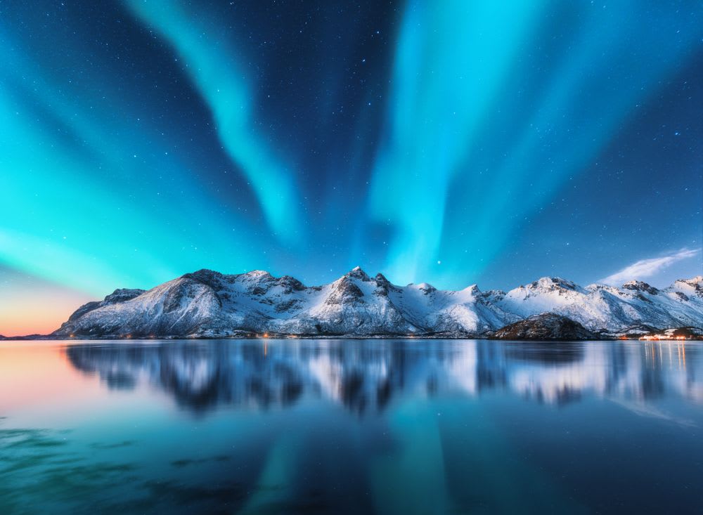 Northern lights dance above snow-covered Icelandic mountains, reflected in a calm fjord under a starry winter sky at twilight