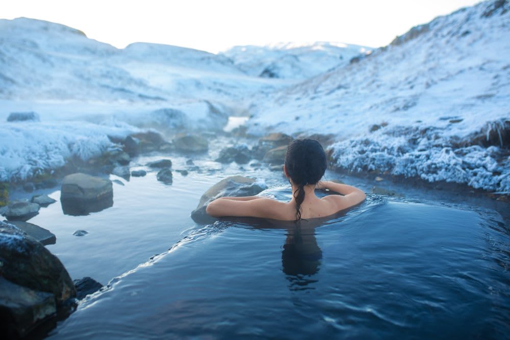 Woman soaking in a steaming Iceland hot spring surrounded by snowy mountains and icy rocks, a relaxing geothermal winter experience