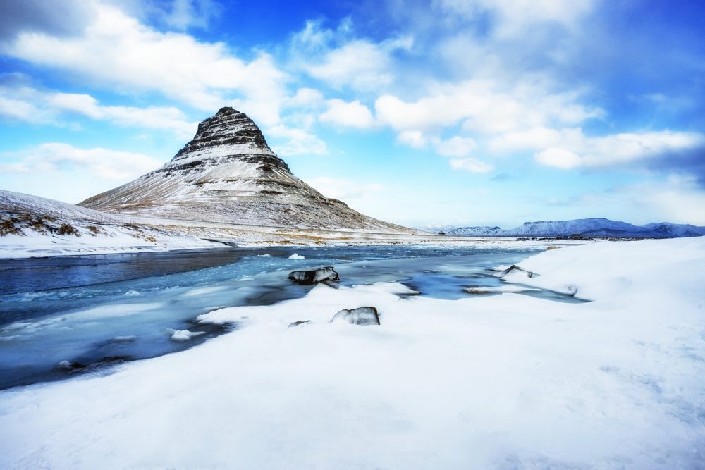 Snow-covered Kirkjufell mountain rises beside an icy lake under blue winter skies on Iceland’s Snæfellsnes Peninsula, perfect for winter travel