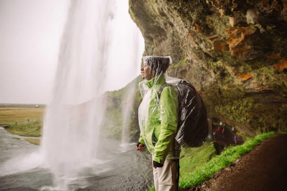 Traveller in rain gear behind Seljalandsfoss waterfall in Iceland, with misty cliffs and river views for UK adventure trips