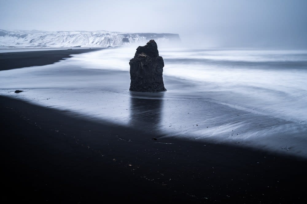 Misty Iceland black sand beach with lone basalt sea stack, snowy cliffs, dark volcanic shore and long-exposure waves along the Atlantic coast
