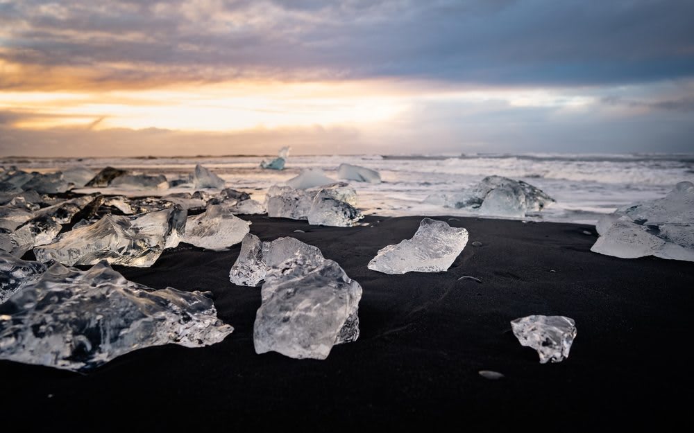 Ice chunks on Diamond Beach in Iceland, scattered across black volcanic sand with Atlantic waves and golden sunset sky near Jokulsarlon