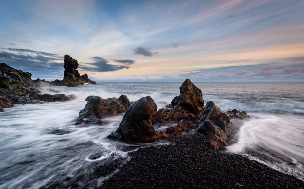Rocky black sand beach in Iceland with volcanic sea stacks, dark lava stones, Atlantic waves and pastel sunset sky along the rugged coast