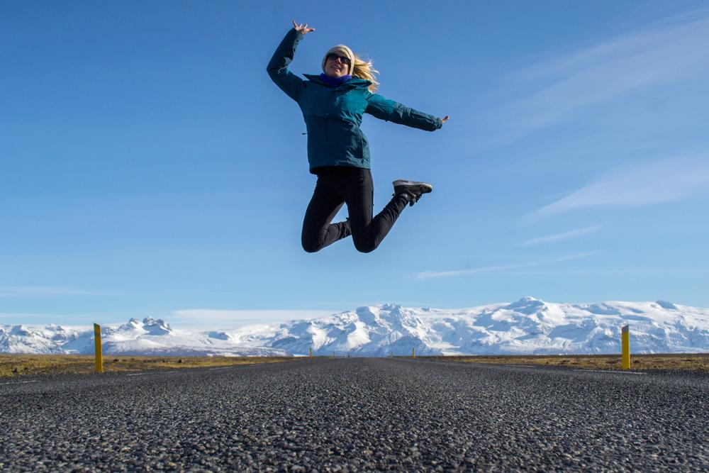 Solo female traveler jumping on Iceland road with snowy glacier mountains under clear blue sky, celebrating freedom and adventure
