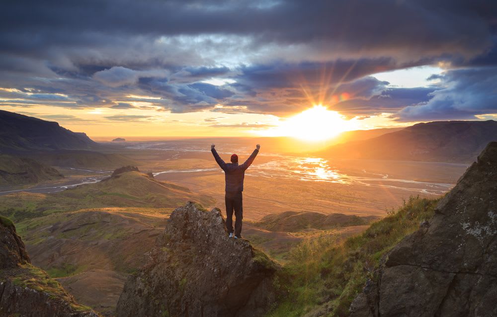 Solo traveler standing on rocky Iceland viewpoint at sunset, arms raised over volcanic valleys, braided rivers and dramatic mountain clouds