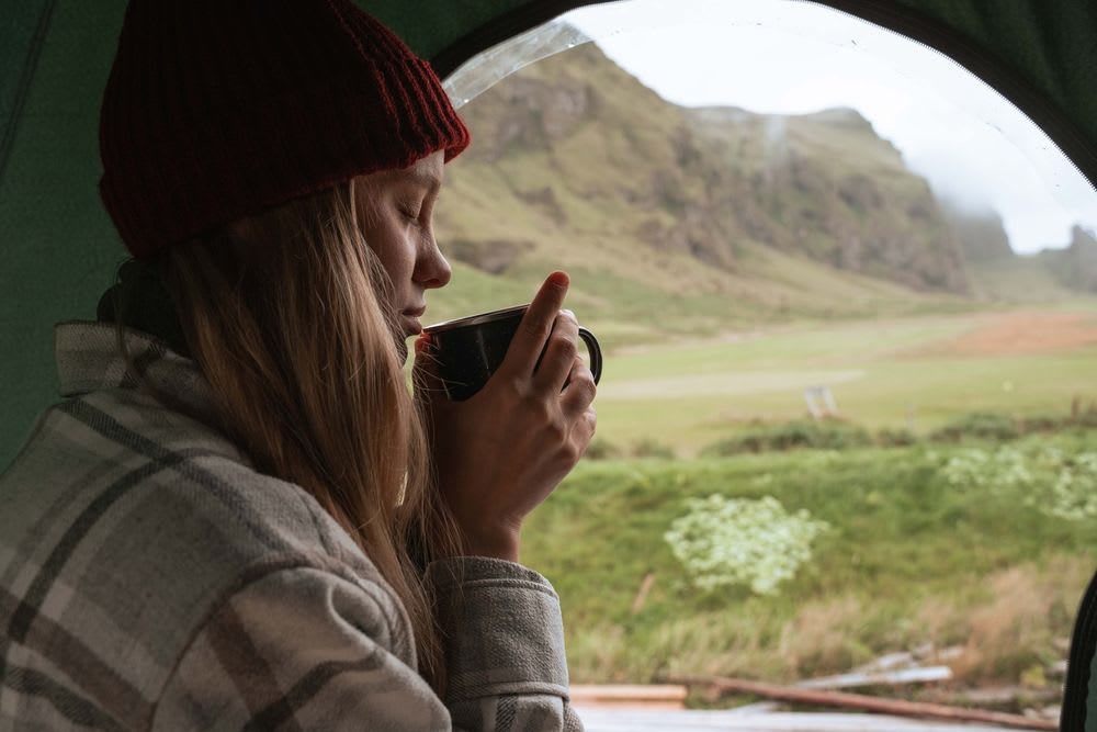 Solo female traveler sipping coffee inside a tent in Iceland, wearing a red beanie and enjoying peaceful mountain views on a cozy camping morning