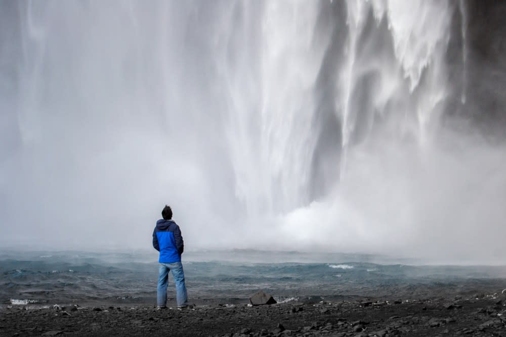 Solo traveler standing before a powerful Iceland waterfall, mist rising over black volcanic rocks and rushing glacial water on an epic solo adventure