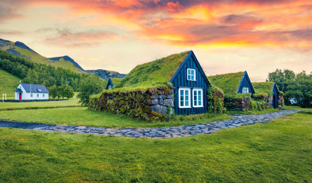 Traditional Icelandic turf houses with grass roofs, stone paths and mountain views under a colorful August sunset in South Iceland