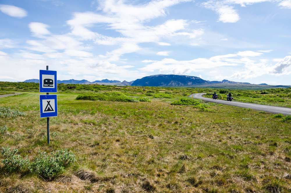 Camping and campervan road signs beside a scenic Iceland highway with motorcyclists, green lava fields and mountains under an August sky