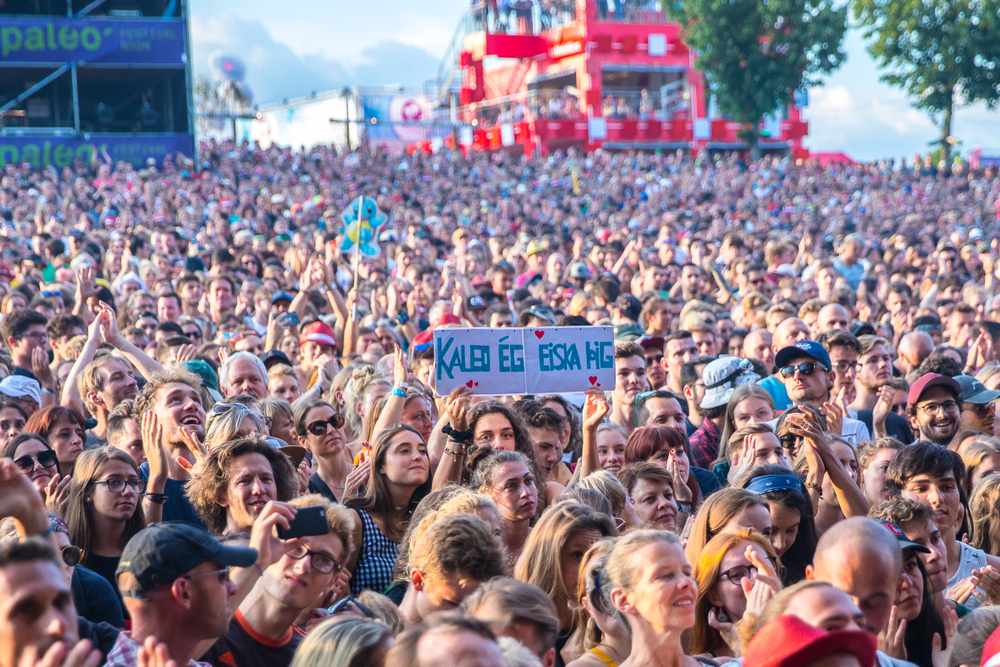 Large crowd cheering at an outdoor summer music festival in Iceland, with a stage, sunny sky and August travel event atmosphere