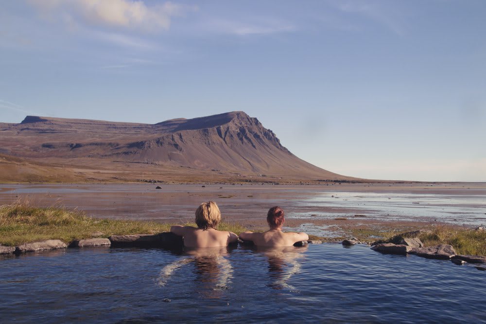 Two travelers relaxing in a natural Iceland hot spring overlooking a fjord, tidal flats and mountains during a calm August evening