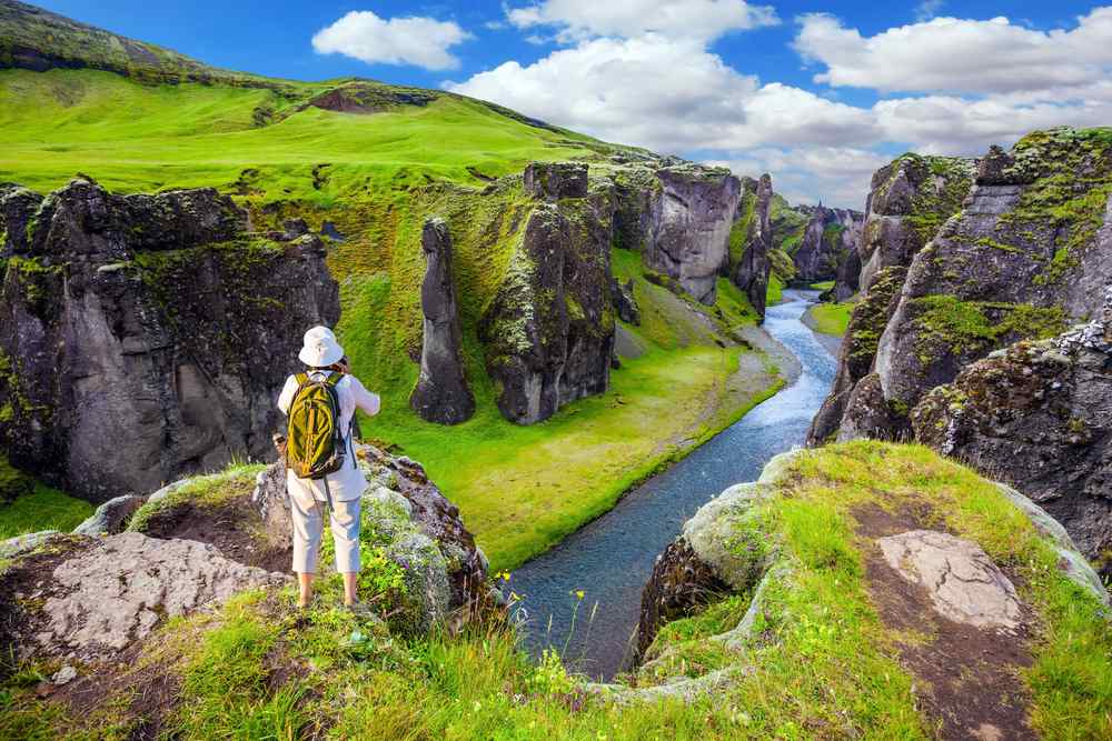 Hiker overlooking Fjaðrárgljúfur canyon in summer, with mossy cliffs, a winding river and vivid green Icelandic landscapes below