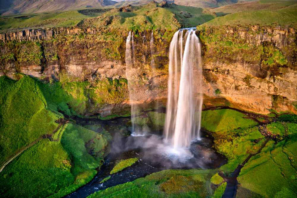 Seljalandsfoss waterfall plunging from green summer cliffs in South Iceland, with mist, basalt rock and lush hiking paths below