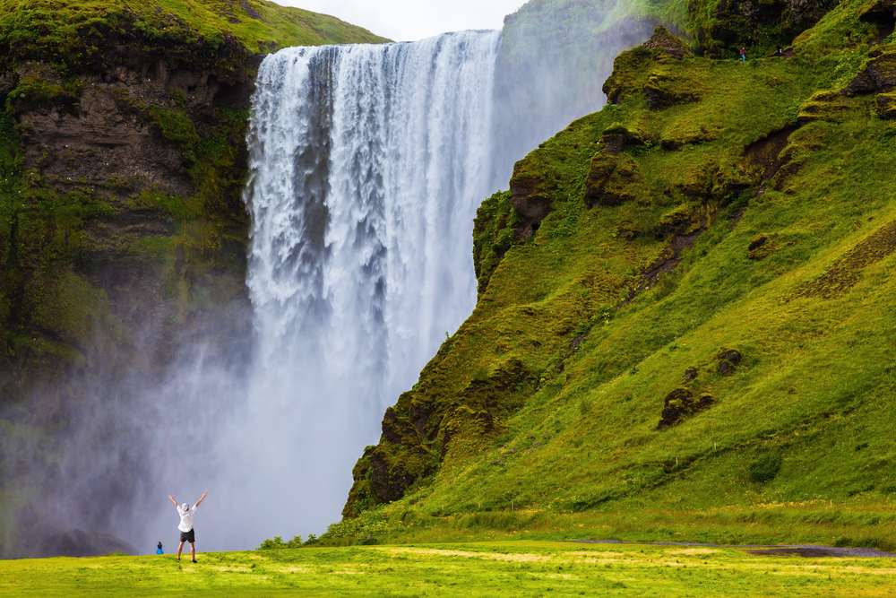 Skógafoss waterfall thundering over mossy cliffs with mist, green hills and a traveler below in South Iceland during July under summer light