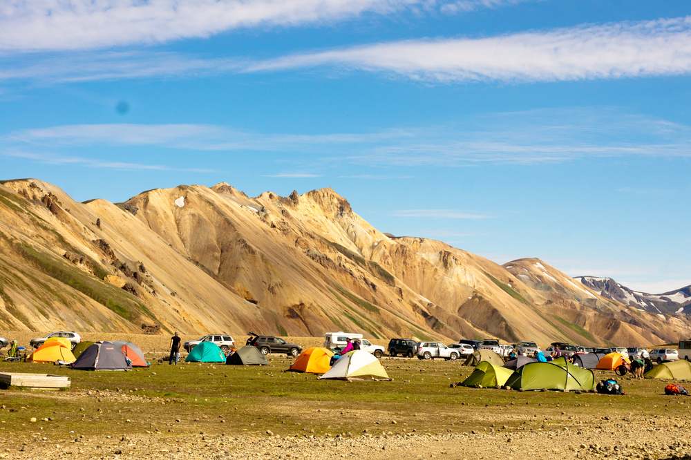 Colorful tents at Landmannalaugar campsite beneath golden rhyolite mountains and blue summer sky in Iceland’s Highlands during July
