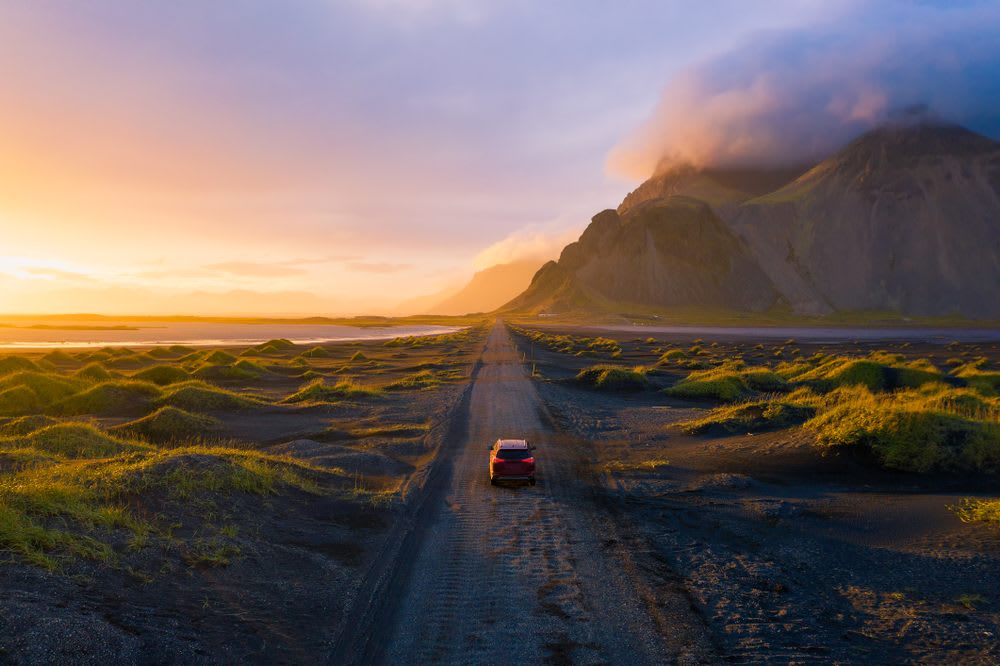 Car driving along a black sand road at Stokksnes, with Vestrahorn mountain, green dunes and golden summer light in Iceland