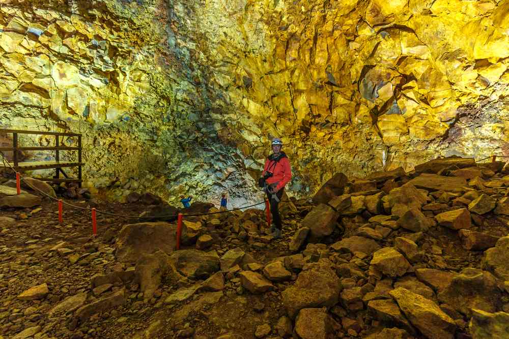 Guide in Raufarhólshellir lava tunnel, surrounded by golden-lit volcanic rock walls and boulders on an Iceland July cave tour