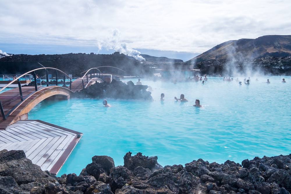 Visitors bathing in the Blue Lagoon’s milky turquoise geothermal waters, with steam, black lava rocks and wooden bridges in Iceland in July