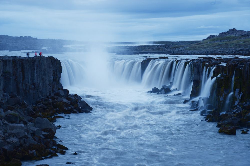 Goðafoss waterfall cascading over dark basalt cliffs into a misty blue river as visitors watch beneath an overcast sky in North Iceland.