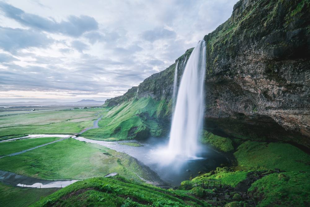 Seljalandsfoss waterfall plunging from a moss-covered cliff above a green valley, with a walking path behind the falls in southern Iceland.