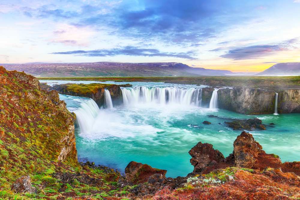 Goðafoss waterfall cascading into a turquoise river at sunset, framed by mossy volcanic rocks and distant mountains in North Iceland.