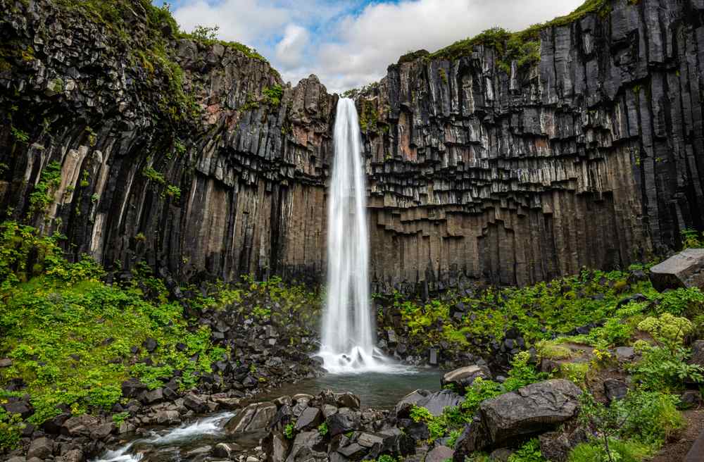 Svartifoss waterfall cascading between black basalt columns into a rocky pool, framed by lush greenery in Vatnajökull National Park, Iceland.