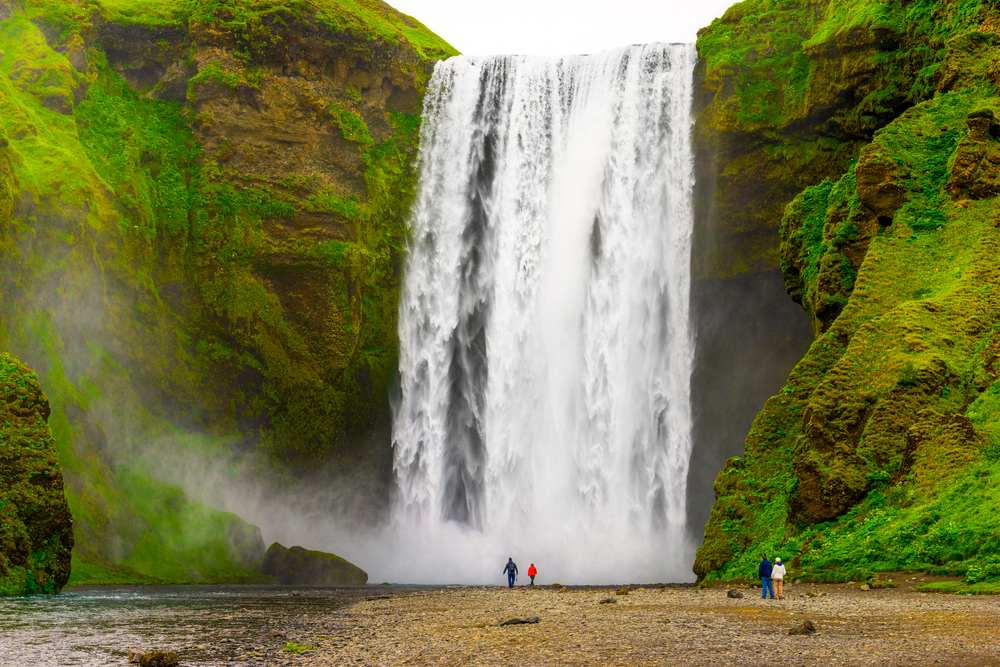Skógafoss waterfall thundering between moss-covered cliffs as visitors stand at its misty base on Iceland’s dramatic South Coast.