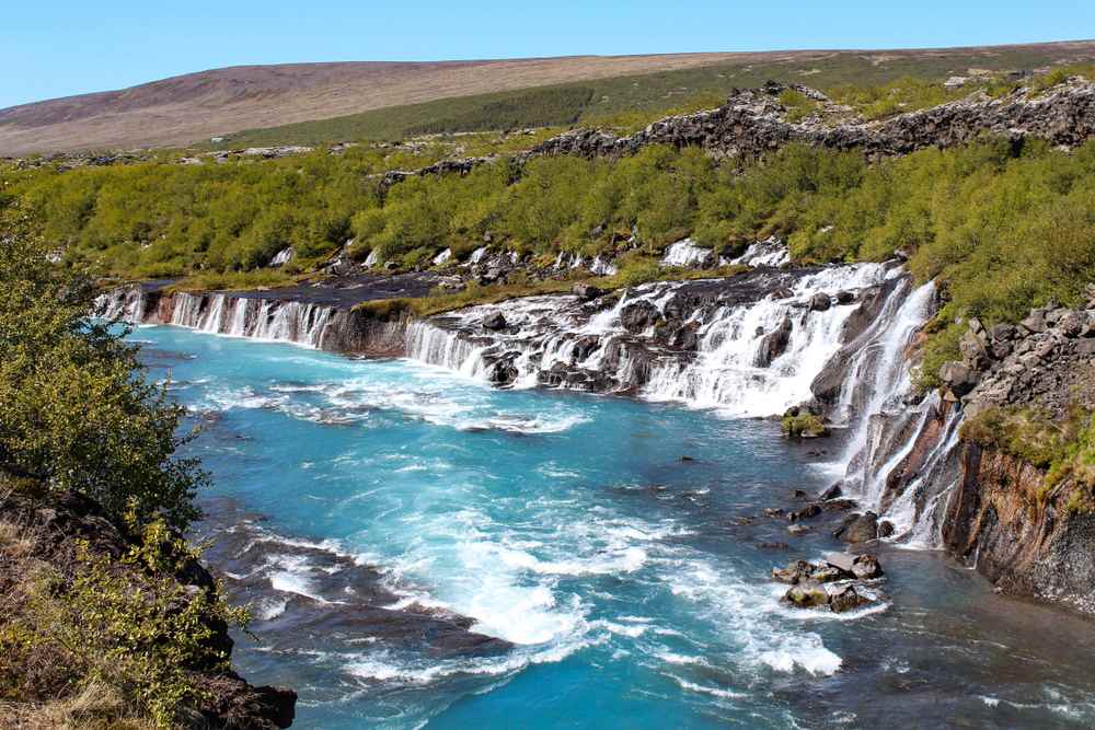 Hraunfossar waterfalls cascading from a lava field into the vivid turquoise Hvítá River beneath clear skies in scenic West Iceland.