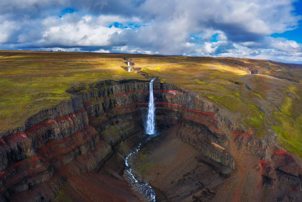 Hengifoss waterfall plunging through red-striped basalt cliffs into a rugged canyon beneath dramatic clouds in scenic East Iceland.
