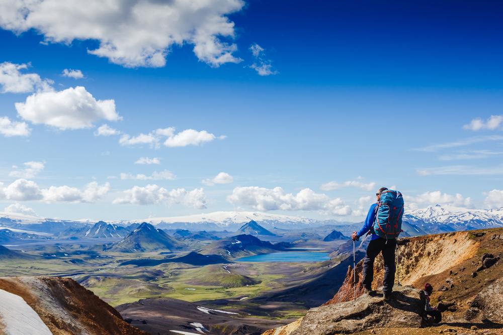 Backpacker with trekking poles overlooks Iceland’s colorful volcanic highlands, turquoise lakes, glaciers and snow-capped peaks beneath a blue sky.