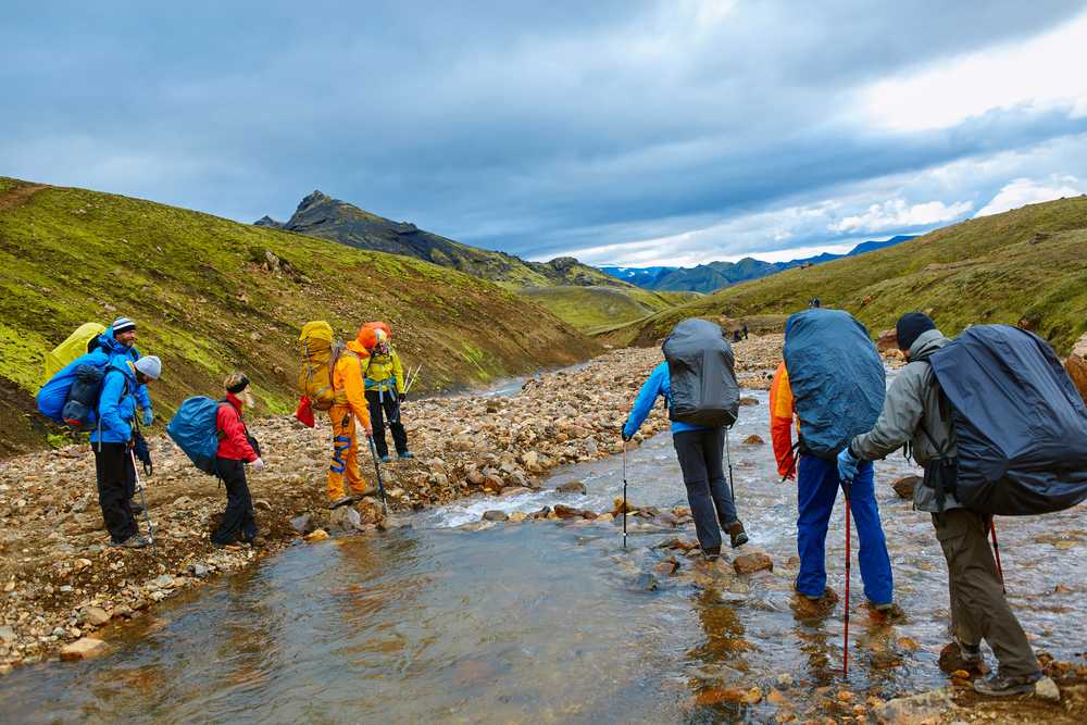 Group of backpackers crossing a rocky stream in Iceland’s remote highlands, surrounded by mossy volcanic hills beneath dramatic storm clouds.