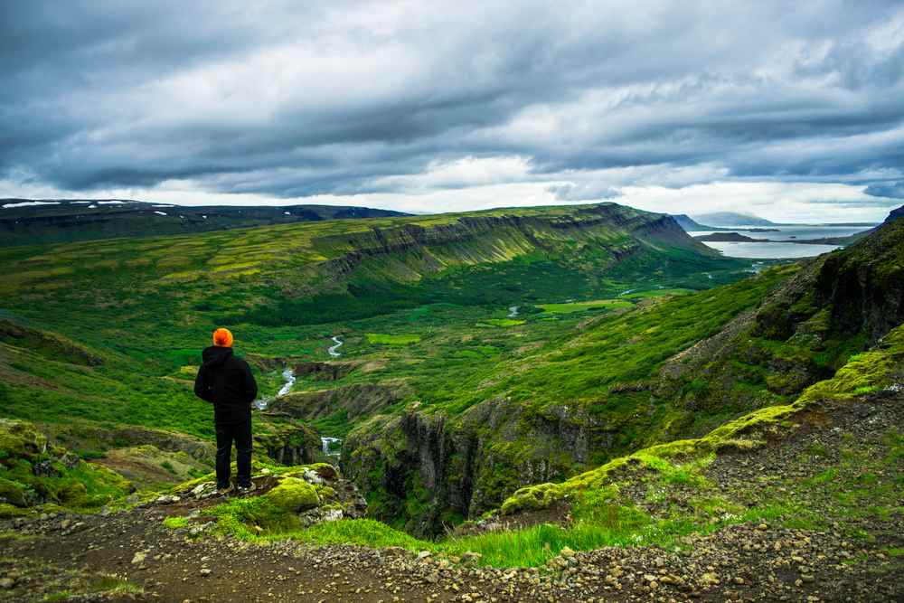 Hiker in an orange hat overlooking a lush Icelandic valley with winding rivers, rugged cliffs and distant fjords beneath dramatic storm clouds.