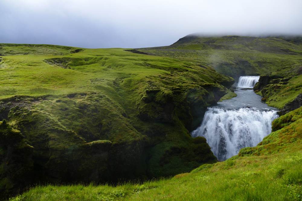 Two-tier waterfall cascading through a misty green Icelandic valley, surrounded by moss-covered cliffs and rolling volcanic highlands.