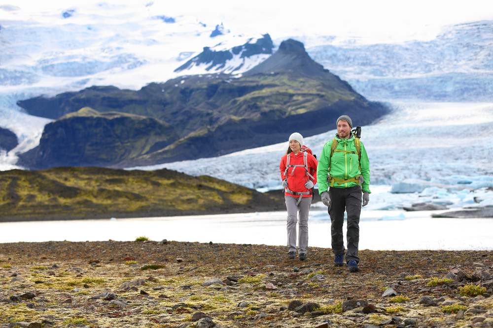 Two backpackers hike across Iceland’s rugged volcanic terrain beside a glacial lagoon, with towering ice fields and dark peaks behind them.