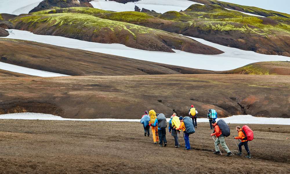 Group of backpackers trekking across Iceland’s barren volcanic highlands, surrounded by moss-covered mountains and lingering snowfields.