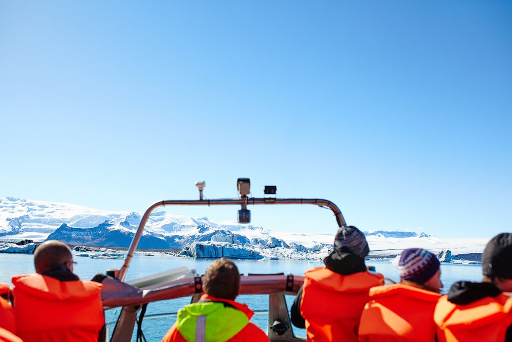 Tourists in orange life jackets take a boat tour across an Icelandic glacial lagoon, surrounded by icebergs and snow-covered mountains.