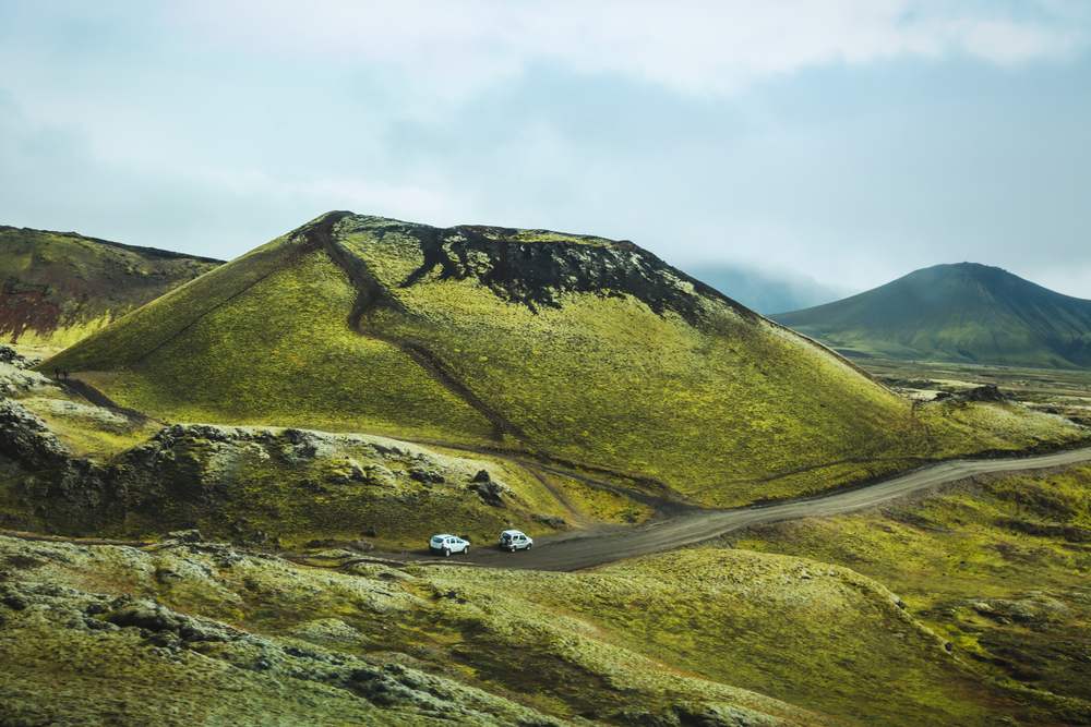 Two white vehicles travel a winding gravel road through moss-covered volcanic mountains in Iceland, showcasing a scenic road trip for US visitors.