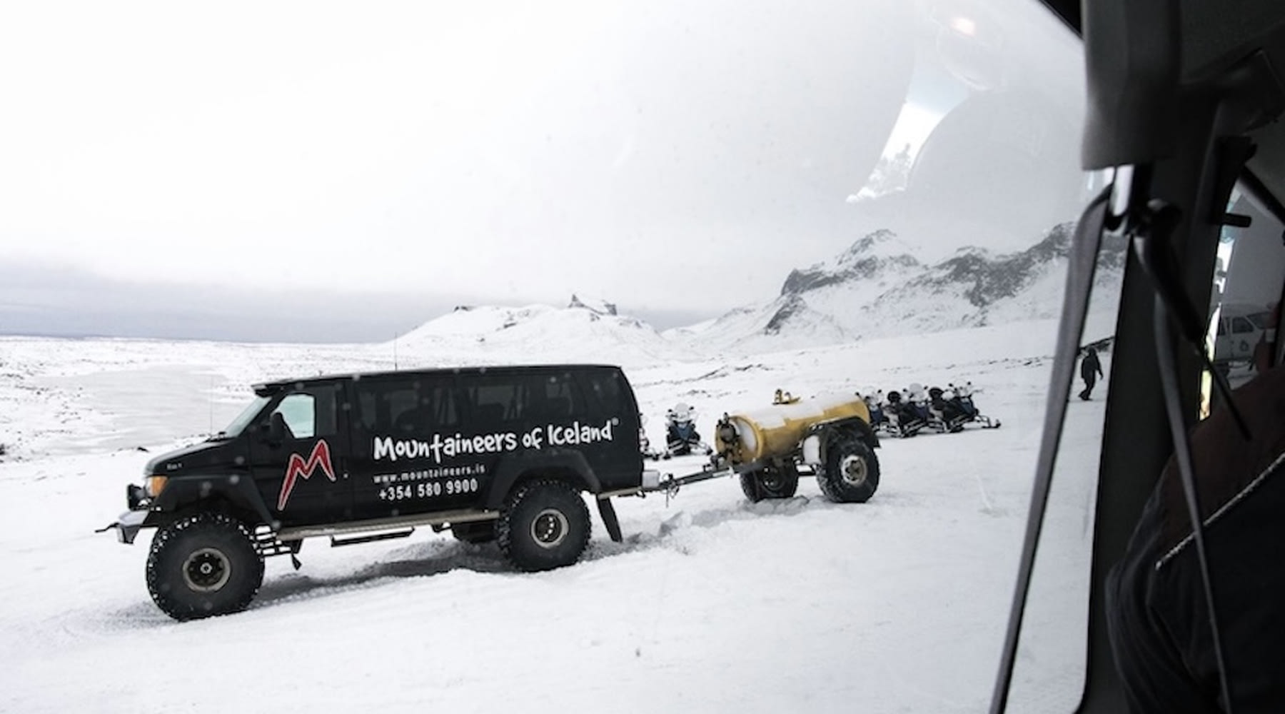 Riesiger Super-Jeep mit einer Reihe von Schneemobilen daneben in einer verschneiten Landschaft.